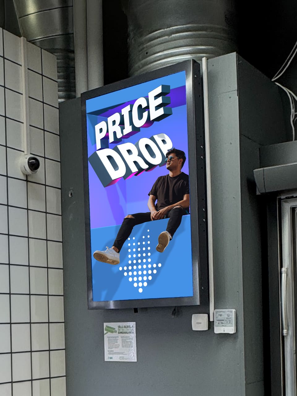 A display mounted on a pillar in a shop. A man sits on the lower edge of a recess in the screen with his legs hanging out over it, under the words PRICE DROP.